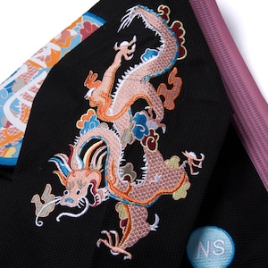 Könnte beinhalten: Schwarzer Stoff mit einem gestickten Drachendesign in Pfirsich-, Orange-, Blau- und Grüntönen. Der Drache ist der Mittelpunkt, mit aufwendigen Details. Ein blau-weißer runder Aufnäher mit den Buchstaben "NS" ist sichtbar.