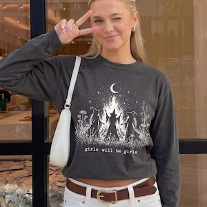 May include: A charcoal gray long-sleeve shirt featuring a white graphic of three figures in pointed hats standing around a campfire under a crescent moon, with the text "girls will be girls" below, paired with a white mini skirt and a white shoulder bag.