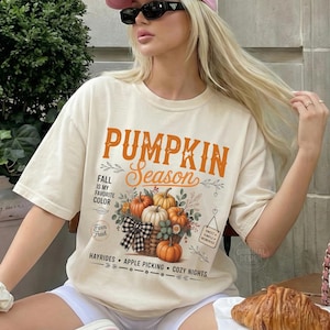 Puede incluir: Una camiseta color marfil con un estampado gráfico que incluye el texto PUMPKIN Season, FALL IS MY FAVORITE COLOR y Farm Fresh. El diseño muestra una cesta de calabazas, un lazo a cuadros blanco y negro y las palabras HAYRIDES, APPLE PICKING, COZY NIGHTS.