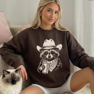 May include: A dark chocolate brown long-sleeve sweatshirt featuring a graphic print of a raccoon wearing a cowboy hat, sunglasses, and a bandana while holding a cup. The text "DARK CHOCOLATE" is printed in the upper right corner of the image.
