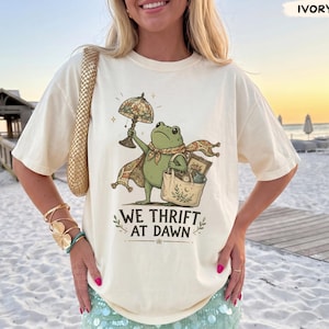 May include: An ivory t-shirt featuring a graphic of a green frog holding a lamp and a tote bag, with the text WE THRIFT AT DAWN printed below. The shirt has short sleeves and a crew neckline, displayed in a casual, loose-fitting style.