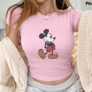 May include: Pink crop top featuring a classic Mickey Mouse graphic. The shirt has short sleeves and a crew neck. The word "PINK" is in the upper right corner. The "Mood Craft Studio" logo is in the lower right corner.
