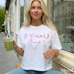 May include: A white short-sleeved t-shirt featuring a pink graphic print of a cowboy boot, a bow, a cowboy hat, and a horseshoe, with the text Cloud 9 written in a cursive script below the icons. The shirt has a crew neckline and a relaxed fit.