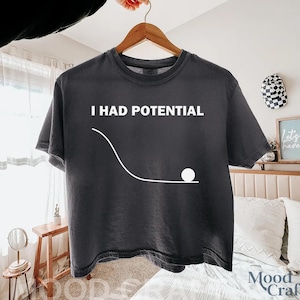 Puede incluir: Camiseta gris oscuro con el texto blanco "I HAD POTENTIAL". Un gráfico blanco representa una bola rodando por una pendiente. La camiseta está colgada en una percha de madera.