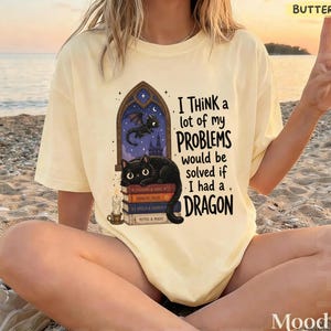 May include: Pale yellow t-shirt with the slogan "I think a lot of my problems would be solved if I had a dragon." The design includes a black cat, a dragon, books, and a candle.