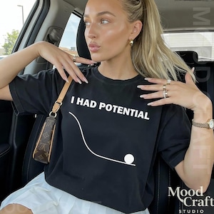 Puede incluir: Camiseta negra con el texto blanco "I HAD POTENTIAL" y un gráfico de una bola rodando por una pendiente. La camiseta la lleva una persona en un coche.