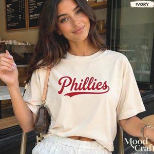 May include: Ivory-colored t-shirt with the word "Phillies" in red script. The shirt is a casual style, likely made of soft fabric. The image shows a person wearing the shirt.