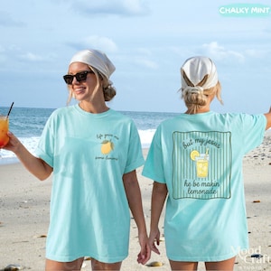 Op de afbeelding: Een licht turquoise t-shirt met een kleine citroenafbeelding en de tekst "life gave me some lemons" op de linkerborst. De achterkant toont een grote afbeelding van een glas limonade met de tekst "but my jesus he be makin lemonade".