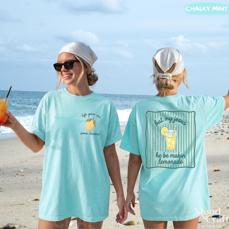 Op de afbeelding: Een licht turquoise t-shirt met een kleine citroenafbeelding en de tekst "life gave me some lemons" op de linkerborst. De achterkant toont een grote afbeelding van een glas limonade met de tekst "but my jesus he be makin lemonade".