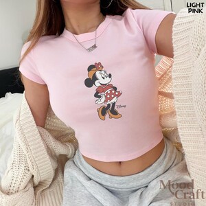 May include: Light pink crop top featuring a Minnie Mouse graphic. The graphic shows Minnie Mouse in a red dress with white polka dots. The top has short sleeves and a crew neck. The word "Disney" is printed below the graphic.