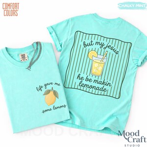 Op de afbeelding: Een krijtmuntkleurig t-shirt met een afbeelding van een gele citroen op de voorkant met de tekst "life gave me some lemons" en een afbeelding van een glas limonade op de achterkant met de tekst "but my jesus he be makin lemonade" over groene strepen.