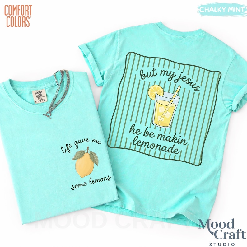 Op de afbeelding: Een krijtmuntkleurig t-shirt met een afbeelding van een gele citroen op de voorkant met de tekst "life gave me some lemons" en een afbeelding van een glas limonade op de achterkant met de tekst "but my jesus he be makin lemonade" over groene strepen.