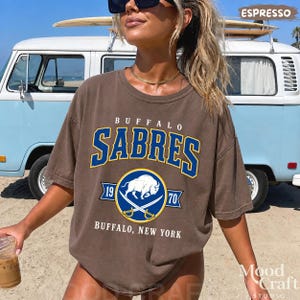 May include: Espresso-colored Buffalo Sabres t-shirt with a blue and white logo. The shirt features the team name in large blue letters, a circular logo with a buffalo, and the text "Buffalo, New York".