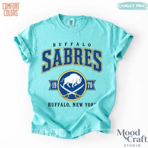 May include: A light blue t-shirt with the text "BUFFALO SABRES" in navy blue and gold. The shirt features a logo with a white buffalo, crossed sabers, and the years 1970 and 1970. The shirt is made by Comfort Colors.