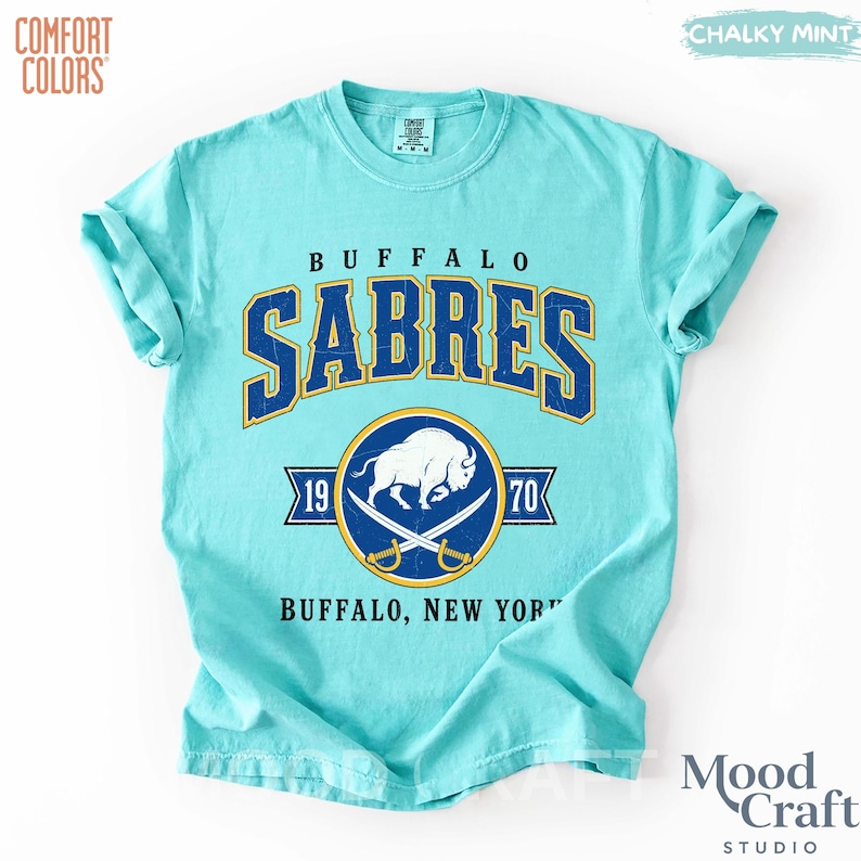 May include: A light blue t-shirt with the text "BUFFALO SABRES" in navy blue and gold. The shirt features a logo with a white buffalo, crossed sabers, and the years 1970 and 1970. The shirt is made by Comfort Colors.