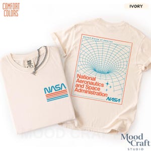 May include: An ivory t-shirt featuring a small NASA logo with blue and orange stripes on the front. The back displays a large graphic of a blue grid representing a black hole, the text National Aeronautics and Space Administration, and NASA.