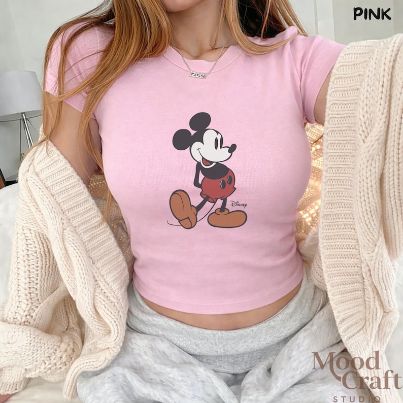May include: Pink crop top featuring a classic Mickey Mouse graphic. The shirt has short sleeves and a crew neck. The word "PINK" is in the upper right corner. The "Mood Craft Studio" logo is in the lower right corner.