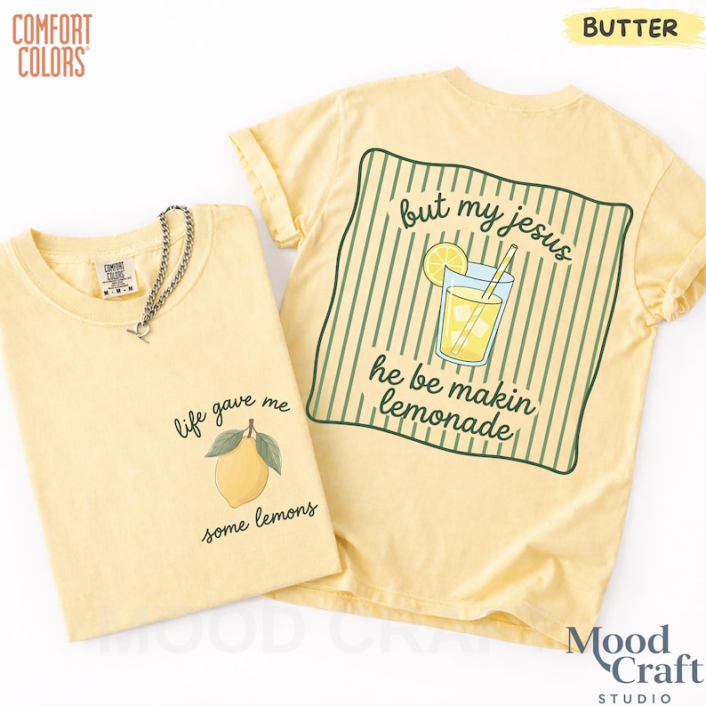 Op de afbeelding: Een botergeel t-shirt met een kleine citroenafbeelding en de tekst 'life gave me some lemons' op de voorkant, en een grote afbeelding op de achterkant met een glas limonade en de tekst 'but my jesus he be makin lemonade'.