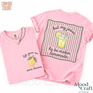 Op de afbeelding: Een lichtroze t-shirt met een kleine gele citroenafbeelding met groene bladeren en de tekst 'life gave me some lemons' op de voorkant, en een groot groen gestreept vierkant met een glas limonade en de tekst 'but my jesus he be makin lemonade' op de achterkant.