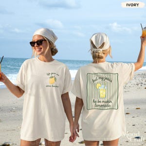 Op de afbeelding: Een ivoorkleurig t-shirt met een kleine citroenafbeelding en de tekst 'life gave me some lemons' op de linkerborst. De achterkant toont een grotere afbeelding van een glas limonade met de tekst 'but my jesus he be makin lemonade'.