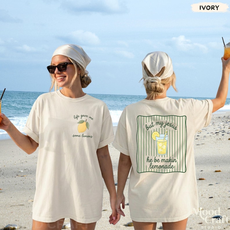 Op de afbeelding: Een ivoorkleurig t-shirt met een kleine citroenafbeelding en de tekst 'life gave me some lemons' op de linkerborst. De achterkant toont een grotere afbeelding van een glas limonade met de tekst 'but my jesus he be makin lemonade'.