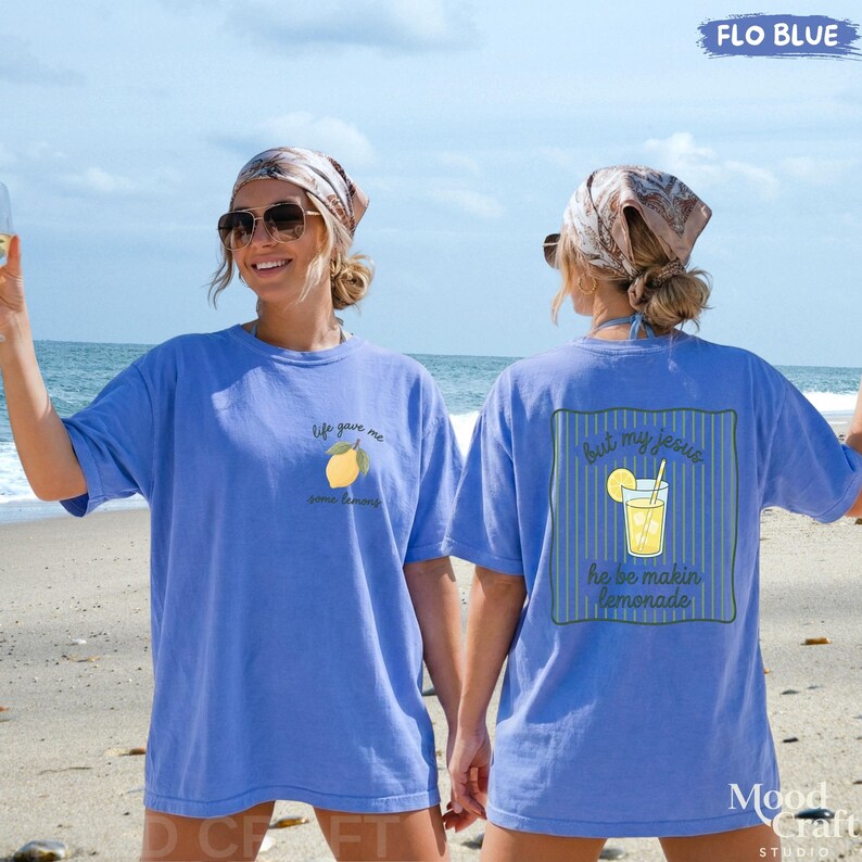 Op de afbeelding: Een korenblauwe t-shirt met een kleine citroenafbeelding en de tekst "life gave me some lemons" op de linkerborst, en een grotere afbeelding op de achterkant met een glas limonade en de tekst "but my jesus he be makin lemonade".