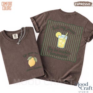 Op de afbeelding: Een donkerbruin t-shirt met korte mouwen, voorzien van een kleine gele citroenafbeelding met de tekst 'life gave me some lemons' op de voorzijde en een groot groen gestreept vierkant met een glas limonade en de tekst 'but my jesus he be makin lemonade' op de achterzijde.