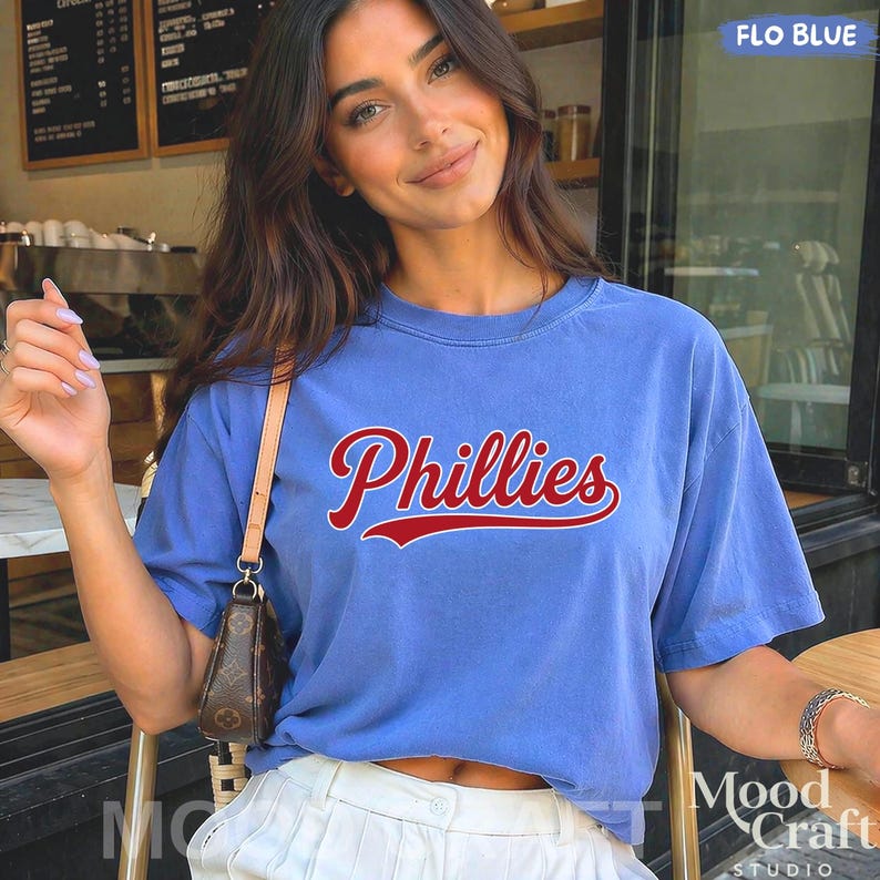 Op de afbeelding: Een lichtblauw t-shirt met het woord "Phillies" in rood script. Het shirt met ronde hals is gemaakt van een zacht materiaal. Het shirt maakt deel uit van een kledingcollectie.