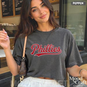 Op de afbeelding: Donkergrijs Phillies T-shirt met rode scriptletters. Het shirt heeft een ronde hals en korte mouwen. De tekst "Phillies" is over de voorkant gebogen. Het shirt wordt gedragen met een witte broek.