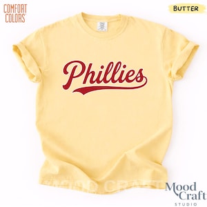 Op de afbeelding: Een geel Comfort Colors T-shirt met het woord "Phillies" in rood script over de voorkant. Het shirt heeft korte mouwen en een ronde hals. Het woord "Butter" staat in de rechterbovenhoek van het shirt.