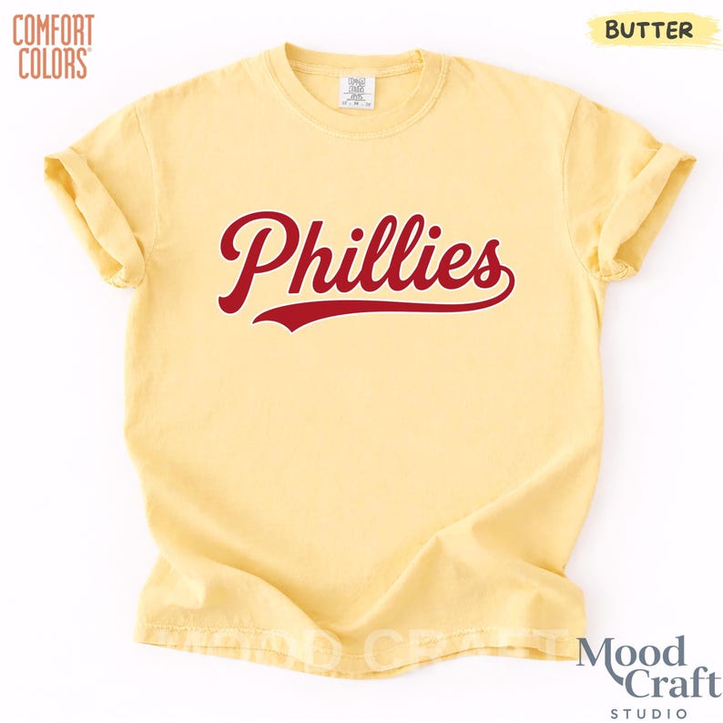 Op de afbeelding: Een geel Comfort Colors T-shirt met het woord "Phillies" in rood script over de voorkant. Het shirt heeft korte mouwen en een ronde hals. Het woord "Butter" staat in de rechterbovenhoek van het shirt.