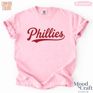 Op de afbeelding: Lichtroze t-shirt met het woord "Phillies" in een rood, cursief lettertype. Het shirt heeft opgerolde mouwen en is gemaakt van een zacht materiaal. De logo's Comfort Colors en Mood Craft Studio zijn zichtbaar.