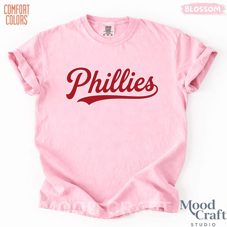Op de afbeelding: Lichtroze t-shirt met het woord "Phillies" in een rood, cursief lettertype. Het shirt heeft opgerolde mouwen en is gemaakt van een zacht materiaal. De logo's Comfort Colors en Mood Craft Studio zijn zichtbaar.