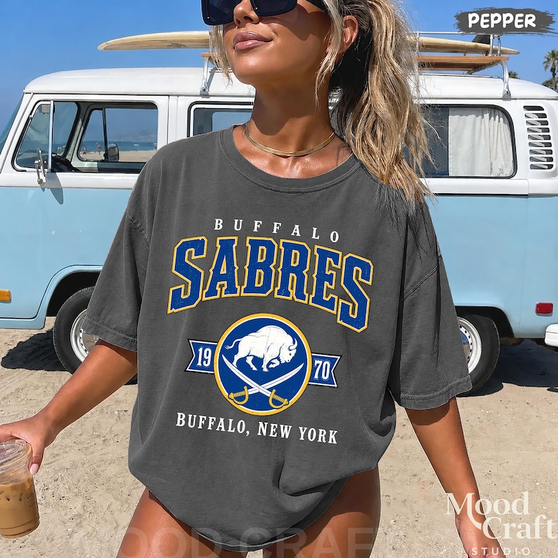 May include: Dark gray t-shirt featuring the Buffalo Sabres logo in blue and gold. The shirt includes the text "BUFFALO SABRES" above a circular emblem with a buffalo and crossed swords. The text "BUFFALO, NEW YORK" is below the emblem.