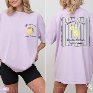 Op de afbeelding: Een lichtpaars t-shirt met een kleine gele citroenafbeelding op de voorkant met de tekst "life gave me some lemons". De achterkant toont een grotere afbeelding van een glas limonade met de tekst "but my jesus he be makin lemonade".