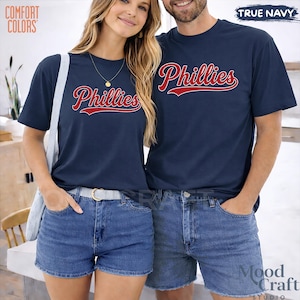 Op de afbeelding: Marineblauwe t-shirts met het woord "Phillies" in rood script. De shirts worden gedragen met denim shorts. De tekst "Comfort Colors" staat in de linkerbovenhoek. De tekst "True Navy" staat in de rechterbovenhoek.
