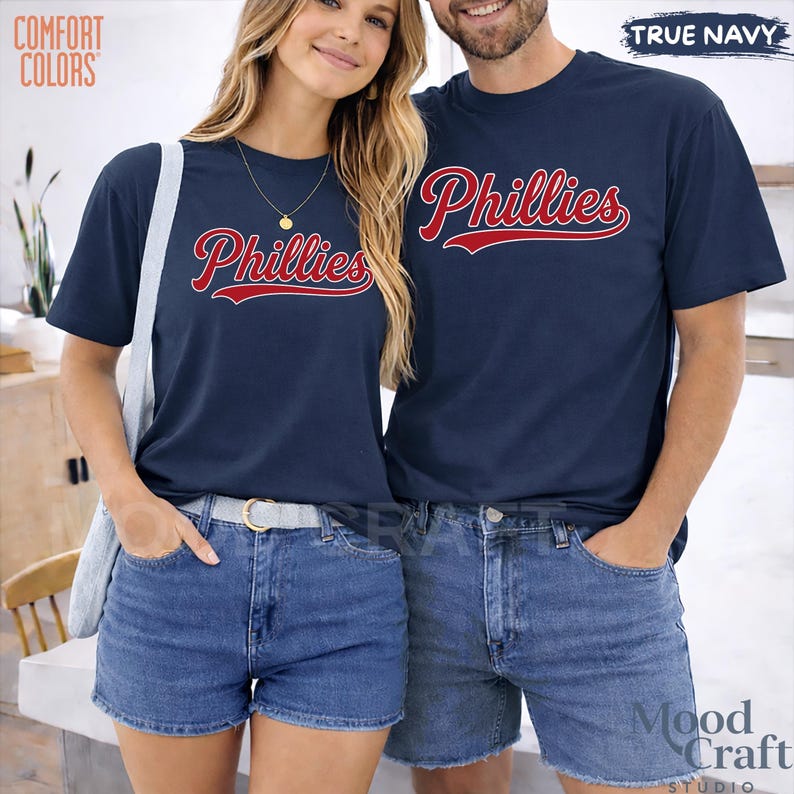 Op de afbeelding: Marineblauwe t-shirts met het woord "Phillies" in rood script. De shirts worden gedragen met denim shorts. De tekst "Comfort Colors" staat in de linkerbovenhoek. De tekst "True Navy" staat in de rechterbovenhoek.