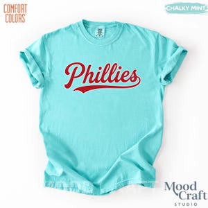 Op de afbeelding: Een lichtblauw t-shirt met het woord "Phillies" in rode letters op de voorkant. Het t-shirt heeft korte mouwen en een ronde hals. De merknaam "Comfort Colors" is linksboven zichtbaar. De tekst "Chalky Mint" staat rechtsboven.