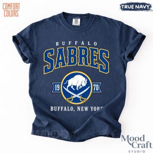 May include: Navy blue t-shirt with "BUFFALO SABRES" in gold and blue lettering. The shirt features a circular emblem with a buffalo and crossed sabers, along with "1970" and "BUFFALO, NEW YORK" text. The shirt is labeled "COMFORT COLORS" and "TRUE NAVY".