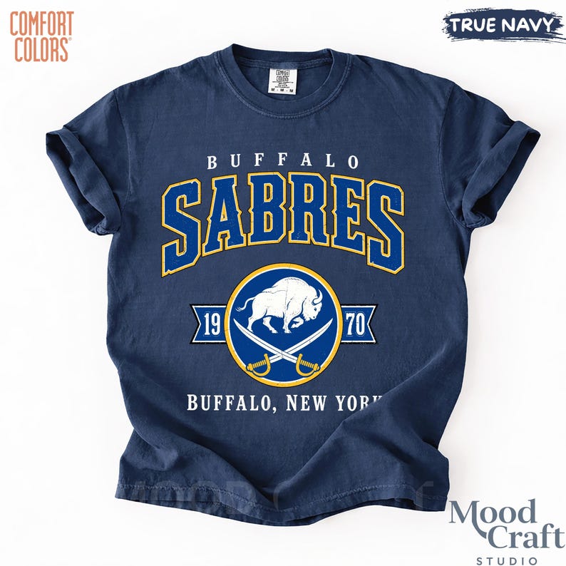 May include: Navy blue t-shirt with "BUFFALO SABRES" in gold and blue lettering. The shirt features a circular emblem with a buffalo and crossed sabers, along with "1970" and "BUFFALO, NEW YORK" text. The shirt is labeled "COMFORT COLORS" and "TRUE NAVY".