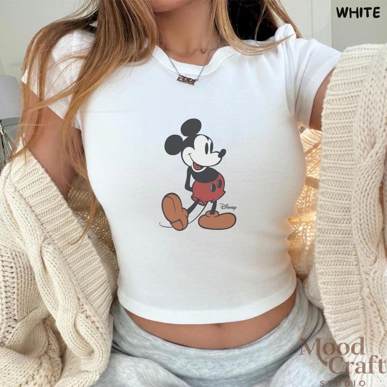 May include: White crop top featuring a classic Mickey Mouse graphic. The cartoon character is depicted in black, red, and brown. The word "Disney" is printed below the character. The top is short-sleeved.