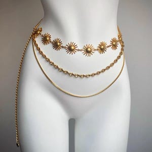 May include: Gold-colored body chain featuring three strands. The top strand has sun-shaped charms. The chain is draped on a white mannequin. The chain is designed to be worn around the waist.
