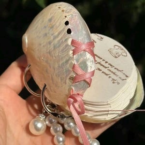 Könnte beinhalten: Eine perlmuttfarbene Muschelschachtel mit rosa Schleifenverschluss. Die Schachtel ist an einem silbernen Schlüsselanhänger mit einem Perlenanhänger und einer Kette aus weißen Perlen befestigt. Die Schachtel enthält kleine, herzförmige Papiere.