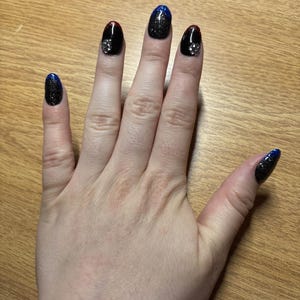May include: Hand with long, oval-shaped fingernails. The nails are painted black with a glittery blue gradient at the tips. Two nails have silver embellishments. The other nails have a red accent at the base.