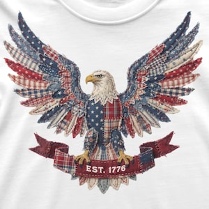 May include: White t-shirt featuring a patriotic eagle design. The eagle has outstretched wings with red, white, and blue patchwork patterns. A banner below reads "EST. 1776". The eagle's head is detailed with a yellow beak and white feathers.