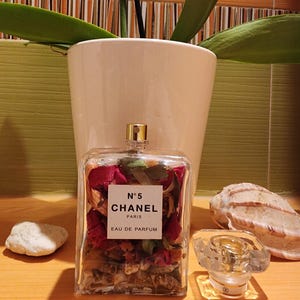 Peut inclure: Flacon rectangulaire de parfum Chanel N°5 rempli de fleurs séchées. Le flacon a un bouchon doré et une étiquette blanche avec du texte noir. Un bouchon de parfum transparent et des coquillages sont à côté.