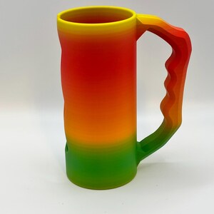 May include: A large, colorful mug with a gradient design. The mug transitions from yellow at the rim to red, orange, and green at the base. The handle mirrors the color scheme, creating a cohesive and vibrant look. The mug is made of a solid material.