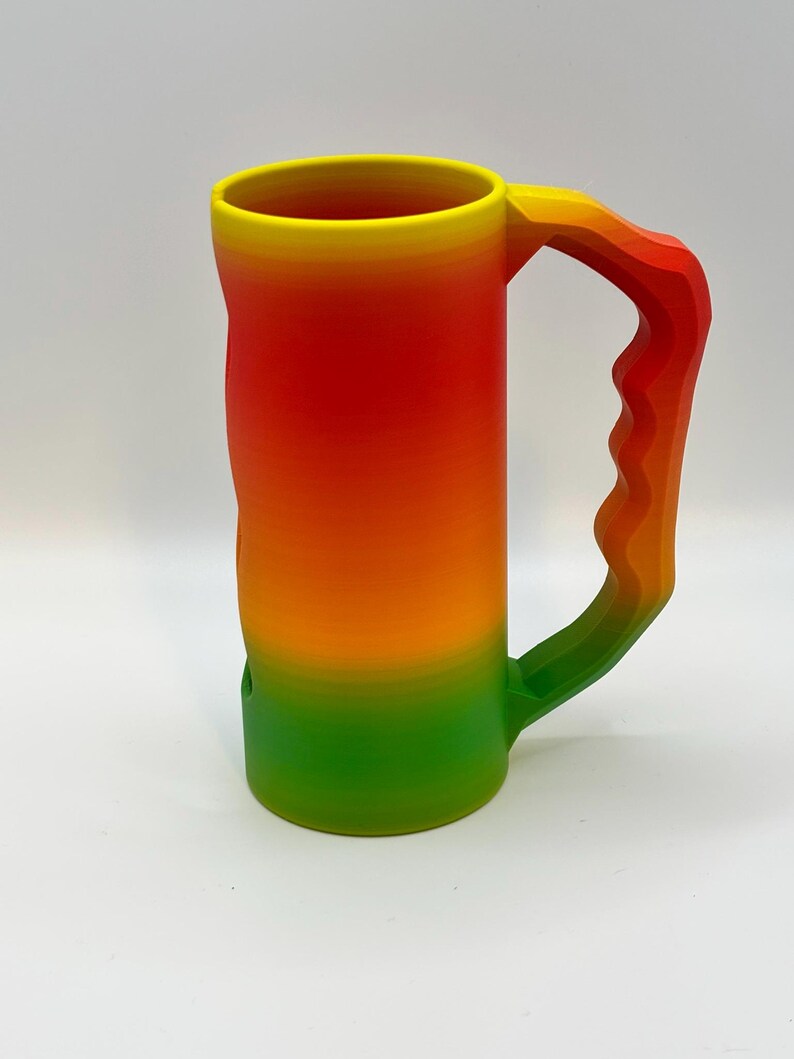 May include: A large, colorful mug with a gradient design. The mug transitions from yellow at the rim to red, orange, and green at the base. The handle mirrors the color scheme, creating a cohesive and vibrant look. The mug is made of a solid material.