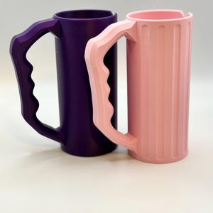 May include: Two cylindrical mugs with large handles. One mug is a deep purple, the other is a light pink. The pink mug has vertical ridges. Both mugs have a unique handle design, and are set against a white background.