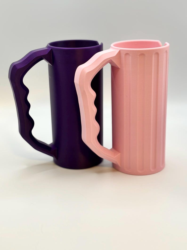 May include: Two cylindrical mugs with large handles. One mug is a deep purple, the other is a light pink. The pink mug has vertical ridges. Both mugs have a unique handle design, and are set against a white background.